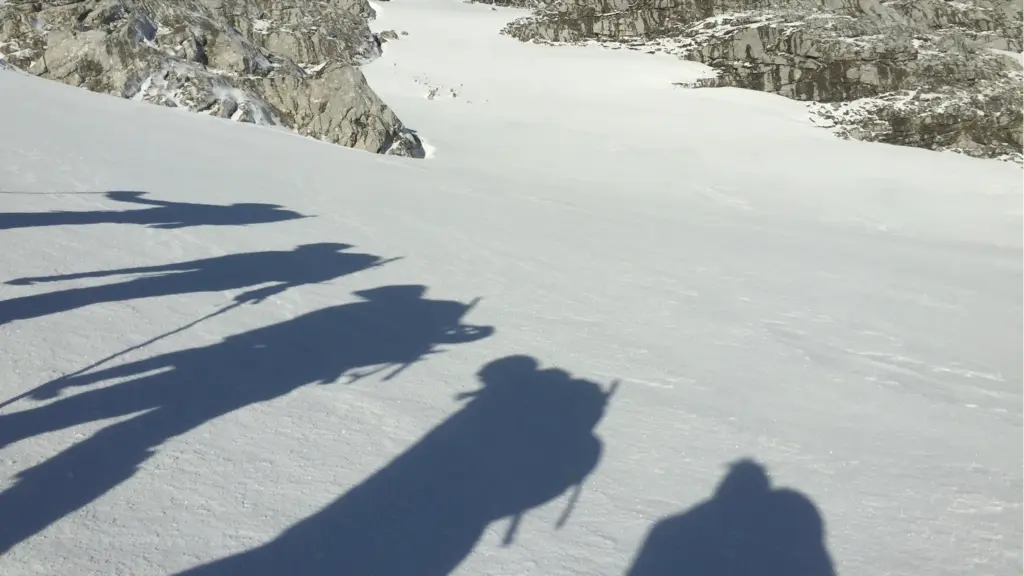 Eine Gruppe Bergsteiger marschiert in einer Reihe über ein Schneefeld, man sieht nur ihre Schatten, die sich lange über den Schnee ziehen.